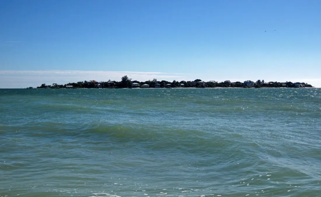 Low barrier island of pale sand and dark vegetation across a shallow blue-green pass under a wide sky.