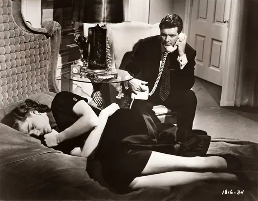 Two actors in a 1956 film still, seated and in conversation.