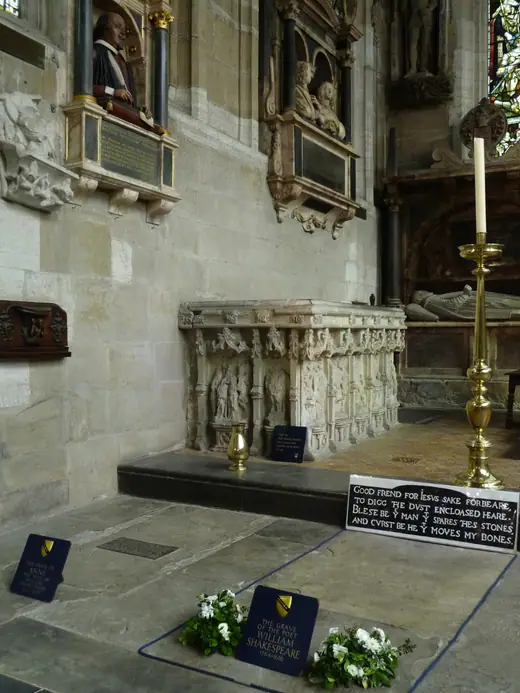 Wall-mounted stone monument with a painted half-length bust of Shakespeare holding a quill, set in an arch with columns.