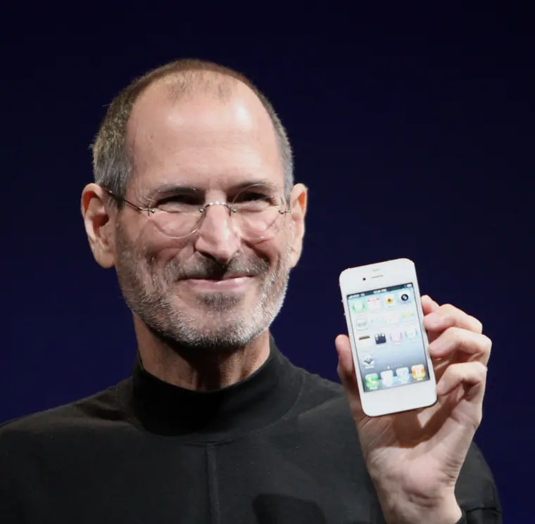 Steve Jobs on stage in a black turtleneck, holding a phone up to the audience.
