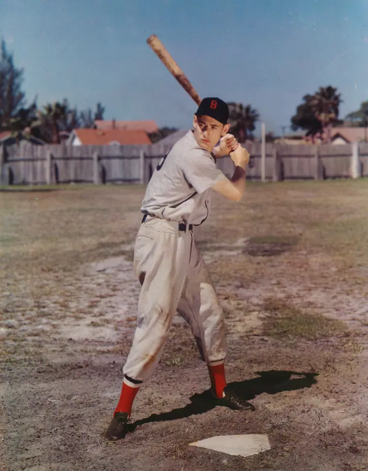 Colour carbro portrait of a young ballplayer in a grey Boston road uniform holding a bat across his shoulder.