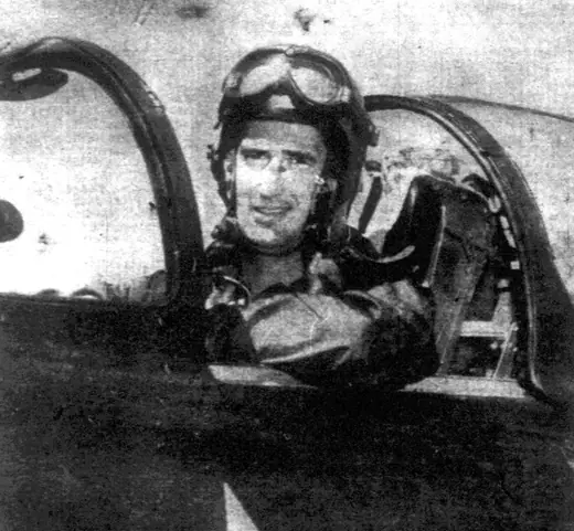 Pilot in flight gear seated in the open cockpit of a jet fighter, looking down at the ground crew.